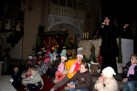 Kobieta ubrana na czarno śpiewa do mikrofonu. Za nią stół liturgiczny i figury. Widać także dzieci siedzące na stopniu ołtarza. 