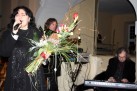 Kobieta z kwiatami w dłoni śpiewa do mikrofonu. W tle dwóch mężczyzn, jeden z nich gra na keyboardzie. 