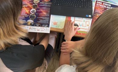 Na zdjęciu dwie młode dziewczyny pracujące na laptopie. Przed nimi plakaty z Dni Ziemi Strzeleckiej. - powiększ