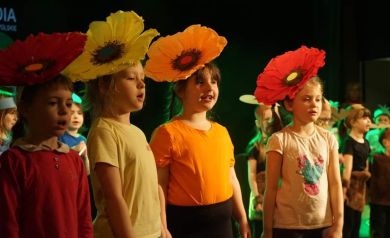 Na zdjęciu widać grupę dzieci na scenie, które biorą udział w przedstawieniu lub koncercie. Czworo dzieci na pierwszym planie ma na głowach duże, kolorowe opaski w kształcie kwiatów. Dzieci śpiewają lub mówią, a w tle widać więcej dzieci ubranych na różne kolory. Scena jest oświetlona zielonym światłem. - powiększ