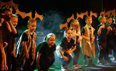 Na zdjęciu jest grupa dzieci na scenie podczas występu. Niektóre dzieci mają na głowach opaski z rogami renifera, a kilka z nich jest ubranych w futrzane kamizelki. W tle jest lekka mgła sceniczna i oświetlenie w ciepłych oraz zielonych kolorach. Dzieci są w różnych pozach, część z nich przykuca. - powiększ