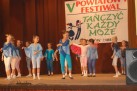 Na zdjęciu widać grupę dzieci występujących na scenie. Dzieci są ubrane w stroje taneczne w odcieniach niebieskiego, białego i różowego, i znajdują się w różnych pozach tanecznych. W tle widoczny jest baner z napisem ,,V POWIATOWY FESTIWAL' oraz „TAŃCZYĆ KAŻDY MOŻE”.