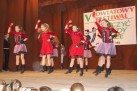 Na zdjęciu widać grupę młodych tancerek na scenie podczas występu. Za nimi znajduje się baner z napisem „V POWIATOWY FESTIWAL TAŃCZYĆ KAŻDY MOŻE”. Dziewczęta są ubrane w czerwono-czarne lub fioletowo-czarne stroje z falbaniastymi spódniczkami i wykonują synchroniczne ruchy taneczne. Widać również część publiczności. 