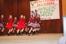 Na zdjęciu widać grupę młodych tancerek na scenie podczas występu. Za nimi znajduje się baner z napisem „V POWIATOWY FESTIWAL TAŃCZYĆ KAŻDY MOŻE”. Dziewczęta są ubrane w czerwono-czarne lub fioletowo-czarne stroje z falbaniastymi spódniczkami i wykonują synchroniczne ruchy taneczne. 