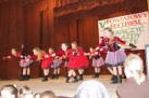 Na zdjęciu widać grupę młodych tancerek na scenie podczas występu. Za nimi znajduje się baner z napisem „V POWIATOWY FESTIWAL TAŃCZYĆ KAŻDY MOŻE”. Dziewczęta są ubrane w czerwono-czarne lub fioletowo-czarne stroje z falbaniastymi spódniczkami i wykonują synchroniczne ruchy taneczne. Widać również część publiczności. 
