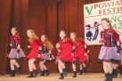 Na zdjęciu widać grupę młodych tancerek na scenie podczas występu. Za nimi znajduje się baner z napisem „V POWIATOWY FESTIWAL TAŃCZYĆ KAŻDY MOŻE”. Dziewczęta są ubrane w czerwono-czarne lub fioletowo-czarne stroje z falbaniastymi spódniczkami i wykonują synchroniczne ruchy taneczne. 
