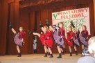 Na zdjęciu widać grupę młodych tancerek na scenie podczas występu. Za nimi znajduje się baner z napisem „V POWIATOWY FESTIWAL TAŃCZYĆ KAŻDY MOŻE”. Dziewczęta są ubrane w czerwono-czarne lub fioletowo-czarne stroje z falbaniastymi spódniczkami i wykonują synchroniczne ruchy taneczne. 