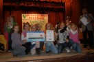 Grupa uśmiechniętych dzieci pozuje na scenie z pucharem i dyplomem podczas V Powiatowego Festiwalu „Tańczyć każdy może”, świętując swoje zwycięstwo.
