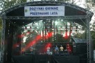 Na zdjęciu widać scenę plenerową z występującym zespołem muzycznym. Scena jest oświetlona reflektorami, z których część emituje czerwone światło. Na scenie znajduje się kilku muzyków grających na instrumentach, takich jak gitara, czy instrumenty dęte, trąbka, saksofon, puzon. Nad sceną wisi baner z napisem 'DOŻYNKI GMINNE POŻEGNANIE LATA'. W tle widoczne są drzewa, co sugeruje, że wydarzenie odbywa się na świeżym powietrzu.