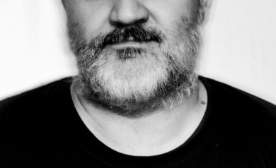 Na zdjęciu widzimy portret mężczyzny w czarno-białej tonacji. Ma on łysą głowę, gęstą brodę i poważny wyraz twarzy. Ubrany jest w ciemną koszulkę z jasnym, okrągłym elementem graficznym widocznym w dolnej części zdjęcia. Tło jest jednolite i jasne, co sprawia, że sylwetka wyraźnie się od niego odcina.