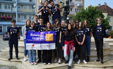 Na zdjęciu znajduje się grupa młodzieży stojąca przed fontanną na miejskim placu. Uczestnicy pozują do wspólnego zdjęcia, większość z nich ma na sobie granatowe koszulki z logo programu „Równać Szanse”. Trzymają baner z nazwą i logotypami projektu. W tle widoczne są budynki mieszkalne oraz Pomnik Myśliwca.  - powiększ
