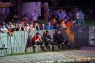 Zdjęcie przedstawia grupę ludzi oglądających wydarzenie plenerowe za barierkami, z przodu siedzi trzech strażaków w mundurach, a za nimi tłum widzów obserwuje scenę. Całość oświetlona jest wieczornym światłem.