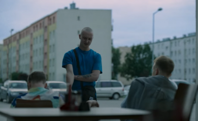 Scena z filmu. Mężczyzna w niebieskiej koszulce prowadzi dialog z kolegami, którzy siedzą na krzesłach.