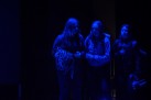 Na zdjęciu widać trzy osoby stojące w ciemnym pomieszczeniu, oświetlone niebieskim światłem. Są częścią sceny teatralnej. Trzymają różne rekwizyty.