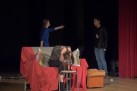 Na zdjęciu trzej aktorzy podczas swojego występu. Jedna z kobiet siedzi na sofie, przed nią stołek, na którym stoi wino i dwa kieliszki do połowy nim zapełnione.