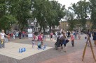 Plac zapełniony dorosłymi oraz dziećmi. Całość otaczają zabudowa miejska i drzewa. Dzieci bawią się m.in. w kręgle. 