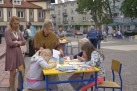 Zdjęcie przedstawia scenę z plenerowego wydarzenia na placu. Główne elementy widoczne na zdjęciu to dwie dziewczynki siedzące przy małym, żółtym stoliku, zajęte rysowaniem lub kolorowaniem. Obok nich stoi kobieta w beżowym swetrze lub ponczo, doglądająca dzieci. Z lewej strony stoi inna kobieta w sukience, z rękami skrzyżowanymi na piersi. W tle widać więcej ludzi, budynki mieszkalne, drzewa i inne stoiska lub namioty. 
