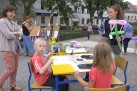Trójka dzieci siedzących przy żółtym stoliku maluje farbami na białych kartkach papieru. Obok stoją dwie dorosłe kobiety. Na drugim planie dzieci i kobieta stojące przy sztalugach. W tle widać inne osoby, zabudowę miejską i roślinność. 