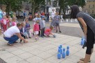 Scena przedstawia grę dzieci w kręgle. Dzieci podzielone są na dwie drużyny. Obok dzieci znajdują się dorośli. W tle zabudowa miejska i roślinność. 