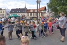 Dzieci idące gęsiego za sobą trzymając się za ramiona. W tle budynki, zielony namiot oraz dorośli. 