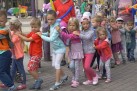 Zdjęcie przedstawia dzieci idące za sobą, trzymają się jeden drugiego za ramiona. W tle widać dorosłych trzymających w dłoniach poskręcane balony. 