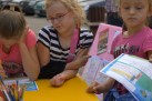 Fotografia przedstawia trzy młode dziewczynki siedzące przy żółtym stole. W centrum znajdują się trzy dziewczynki. Jedna z lewej ma różową koszulkę i pochyla głowę nad stołem. Dziewczynka w środku ma czarno-białą koszulkę w paski, różowe okulary i opiera łokcie na stole. Dziewczynka z prawej strony, w różowej koszulce, trzyma kartkę papieru z nadrukami i ołówek lub długopis. Na stole leżą kartki papieru oraz pojemnik z kredkami. Tło sugeruje, że wydarzenie odbywa się na zewnątrz lub w otwartej przestrzeni, z niewyraźnymi elementami takimi jak samochody i inne osoby w oddali.
