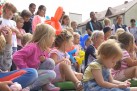 Na pierwszym planie grupa dzieci z kolorowymi balonikami w dłoniach. Jedna z dziewczynek ma założoną kolorową maskę na twarzy. Z tyłu widać osoby dorosłe. W tle znajdują się budynki i widać część zielonego namiotu. 