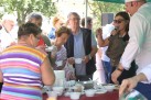 Na zdjęciu widoczne jest zgromadzenie ludzi na świeżym powietrzu, podczas pikniku familijnego. Główne elementy widoczne na zdjęciu to grupa osób w różnym wieku stoi wokół stołu. Kilka osób, w tym jedna w stroju szefa kuchni i kobieta z mikrofonem, wydaje coś (prawdopodobnie zupę lub inny posiłek) do małych, białych kubków. Na stole znajdują się białe kubki, miski, serwetki i inne przybory kuchenne. W tle widać zieleń (drzewa lub krzewy).
