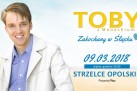 Plakat zapowiada koncert Toby’ego z Monachium zatytułowany „Zakochany w Śląsku”, który odbył się 9 marca 2018 roku o godzinie 19:00 w Strzelcach Opolskich. Po lewej stronie widnieje uśmiechnięty mężczyzna w jasnej marynarce i niebieskiej koszuli, a po prawej umieszczono informacje o wydarzeniu. Tło jest jasne i pastelowe, co nadaje całości lekkiego i pogodnego charakteru.