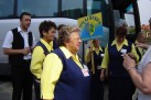 Zespół ,,Viva la Musica' przy autobusie. Kobiety ubrane są w żółto granatowe stroje. Mężczyzna stojący obok nich ma na sobie ciemne spodnie, kamizelkę i białą koszulę. Jedna z kobiet trzyma tablicę z napisem ,,Viva la Musica Strzelce Opolskie'. 