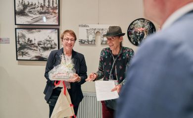 Dwie kobiet podczas wernisażu, kobieta po lewej trzyma na rękach upominek, kobieta po prawej w kapeluszu w przyjaznej atmosferze 