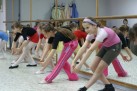 Na zdjęciu widać grupę dziewczynek ćwiczących w sali tanecznej przed lustrem — wykonują one skłony. Wszystkie mają na sobie stroje sportowe lub baletowe.